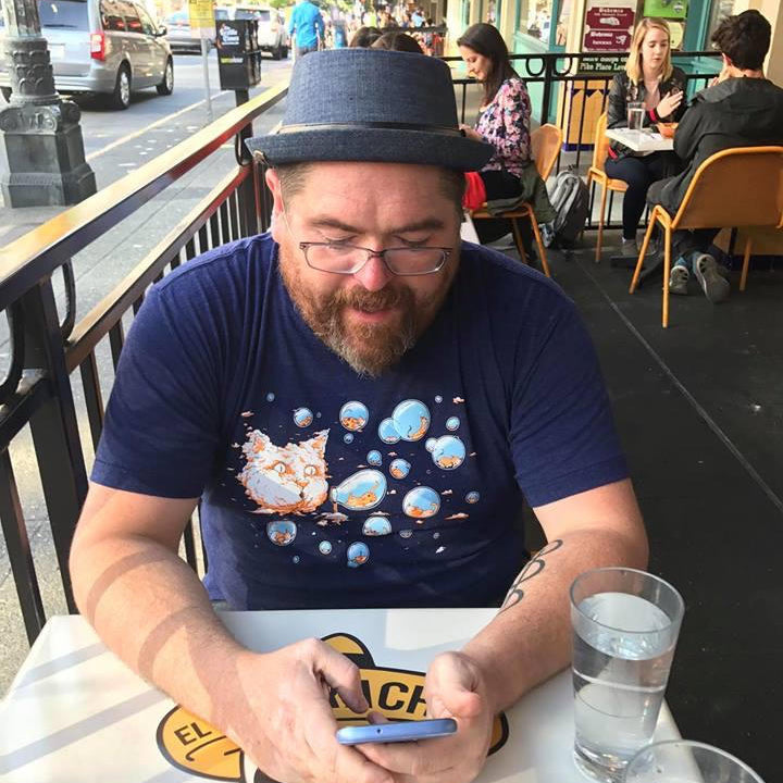 Man sitting at an outdoor cafe using a phone, with a city street in the background. He is wearing a hat and a bubble cat shirt.
