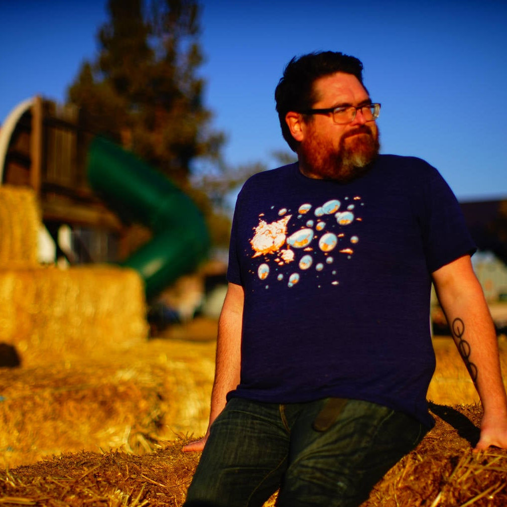 Man sitting on hay bales with a playground in the background. He is wearing a bubble cat shirt. It has a big cat head shaped like a cloud and the cloud cat is blowing bubbles. Inside the bubbles are tiny kittens. 
