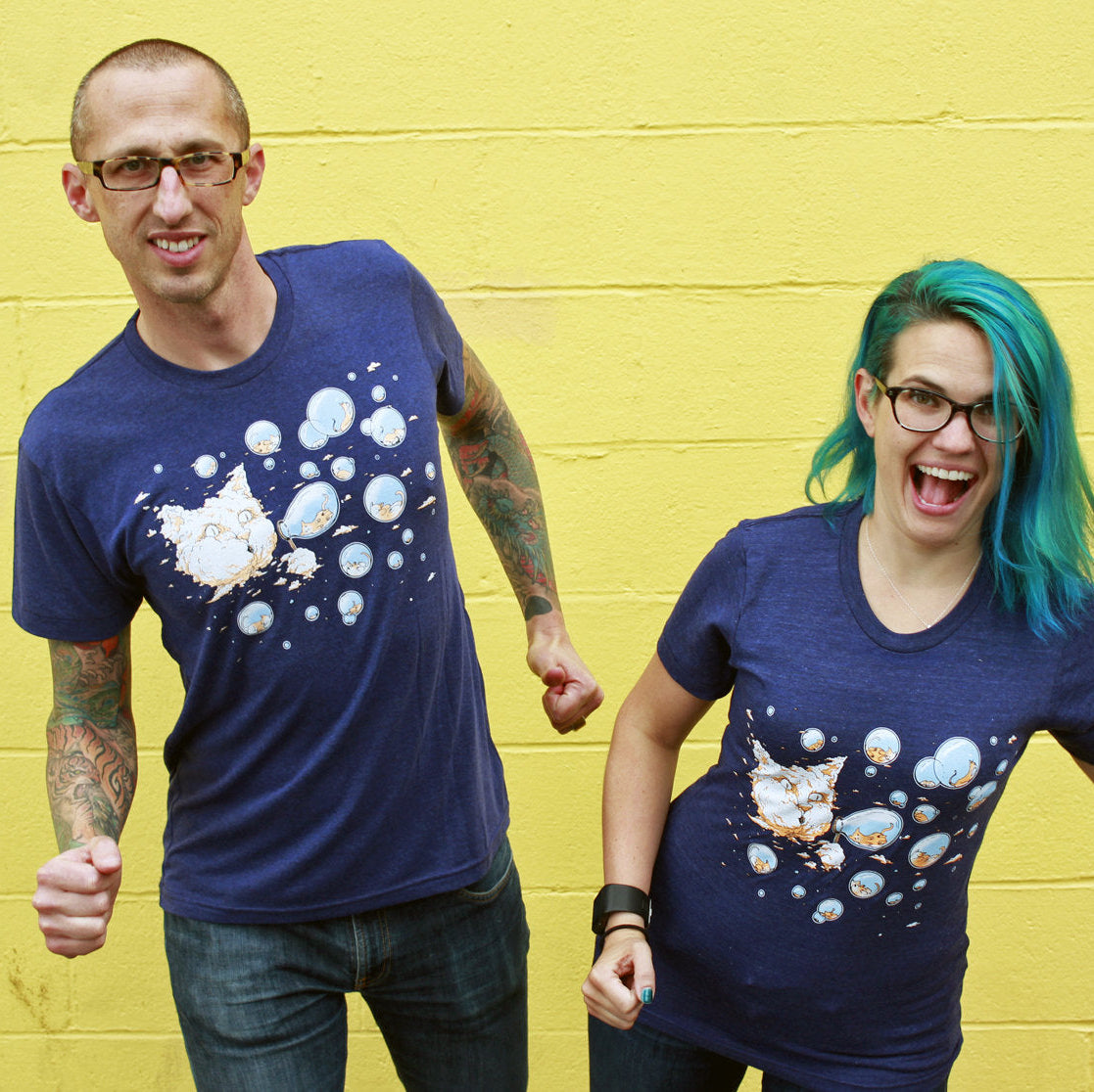 A man and a woman standing next to each other, both wearing a blue t-shirt with a cloud and bubble design. The man is wearing glasses. The woman has blue hair and is also wearing glasses.