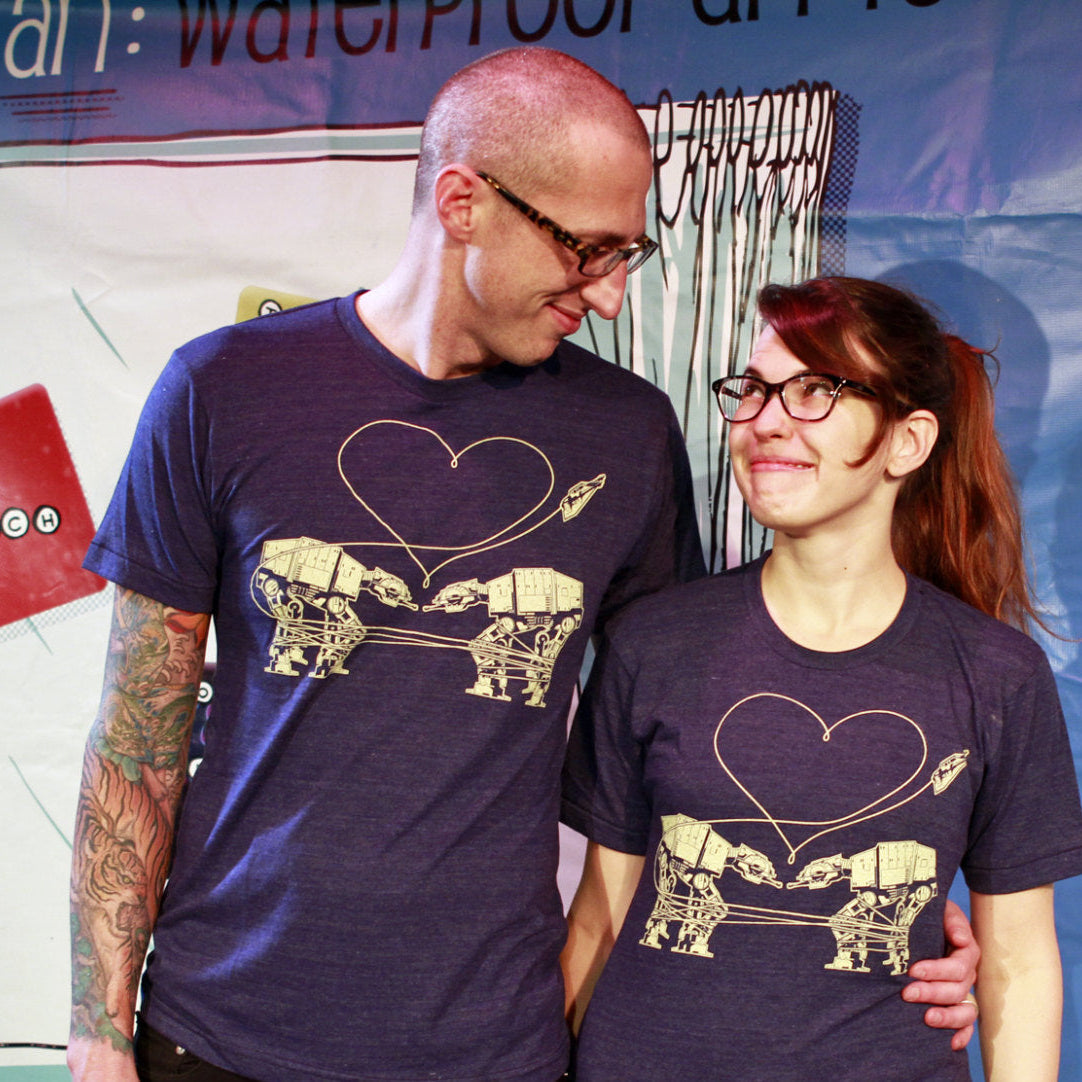 Two people wearing matching t-shirts with two AT-ATs falling in love. 