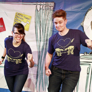 Two people wearing matching heather blue t-shirts with two AT-Ats falling in love, standing in front of a colorful background.