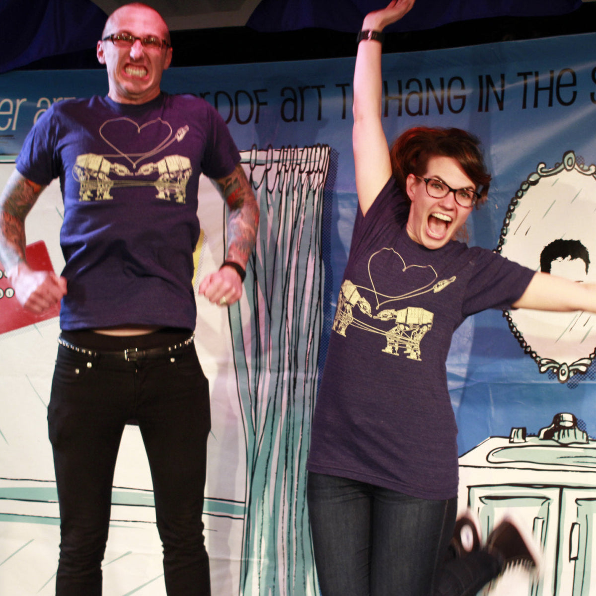 Two people wearing matching heather blue t-shirts with a screenprinted design of two AT-ATs falling in love, standing in front of a colorful backdrop.