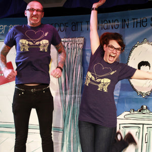 Two people wearing matching heather blue t-shirts with a screenprinted design of two AT-ATs falling in love, standing in front of a colorful backdrop.