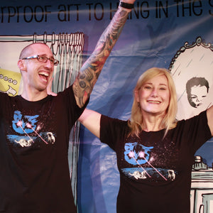 Two people with tattoos and black t-shirts standing in front of a colorful mural.