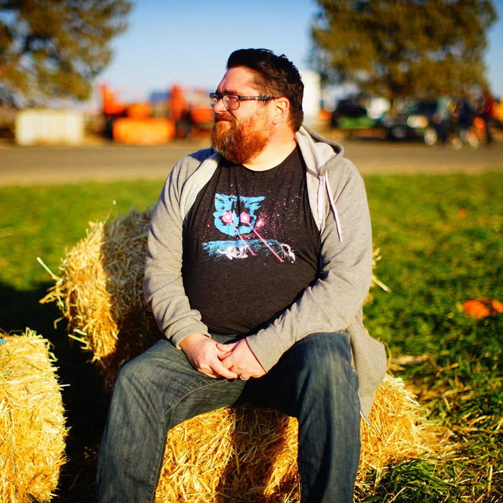A person sitting on a bale of hay, wearing a gray hoodie and a black tshirt with a cat head shooting lasers out of his eyes. 