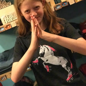 Person wearing a black t-shirt with a roller skating unicorn design, standing in front of a wooden shelf.