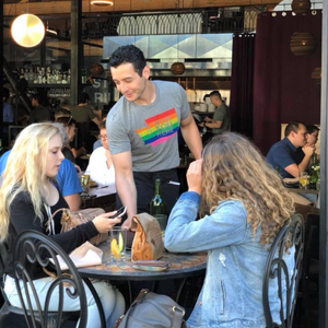 Man in a gray t-shirt with a rainbow design interacting with two women at an outdoor cafe.