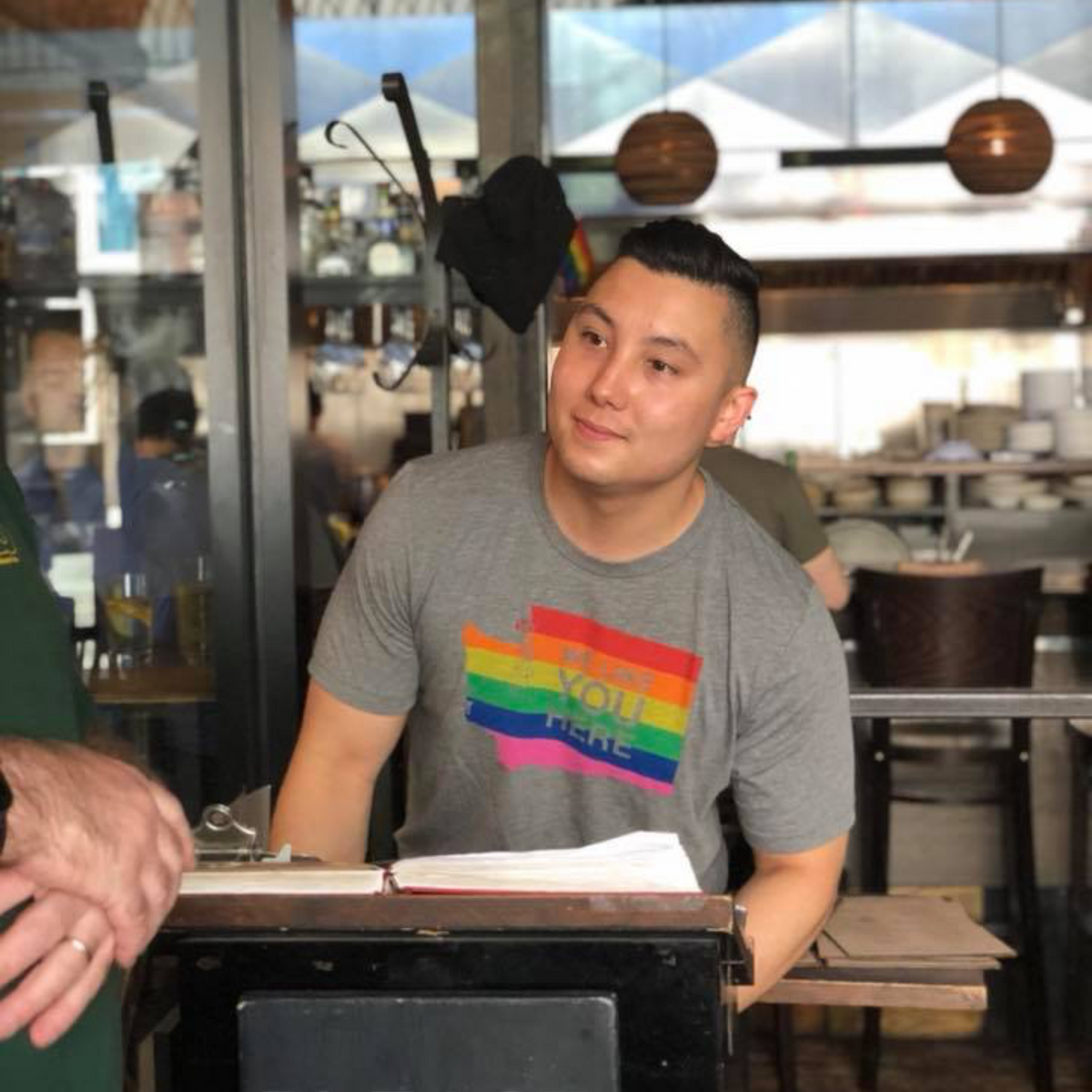 Person wearing a gray t-shirt with a rainbow design sitting at a counter in a restaurant.