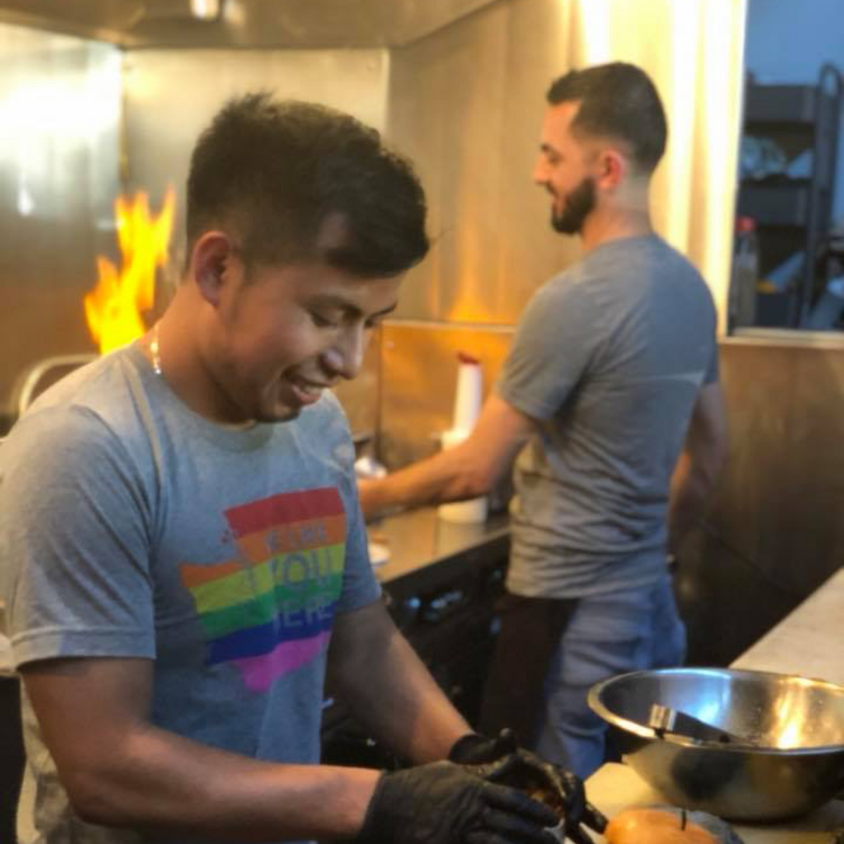 Two men preparing food in a kitchen with flames in the background
