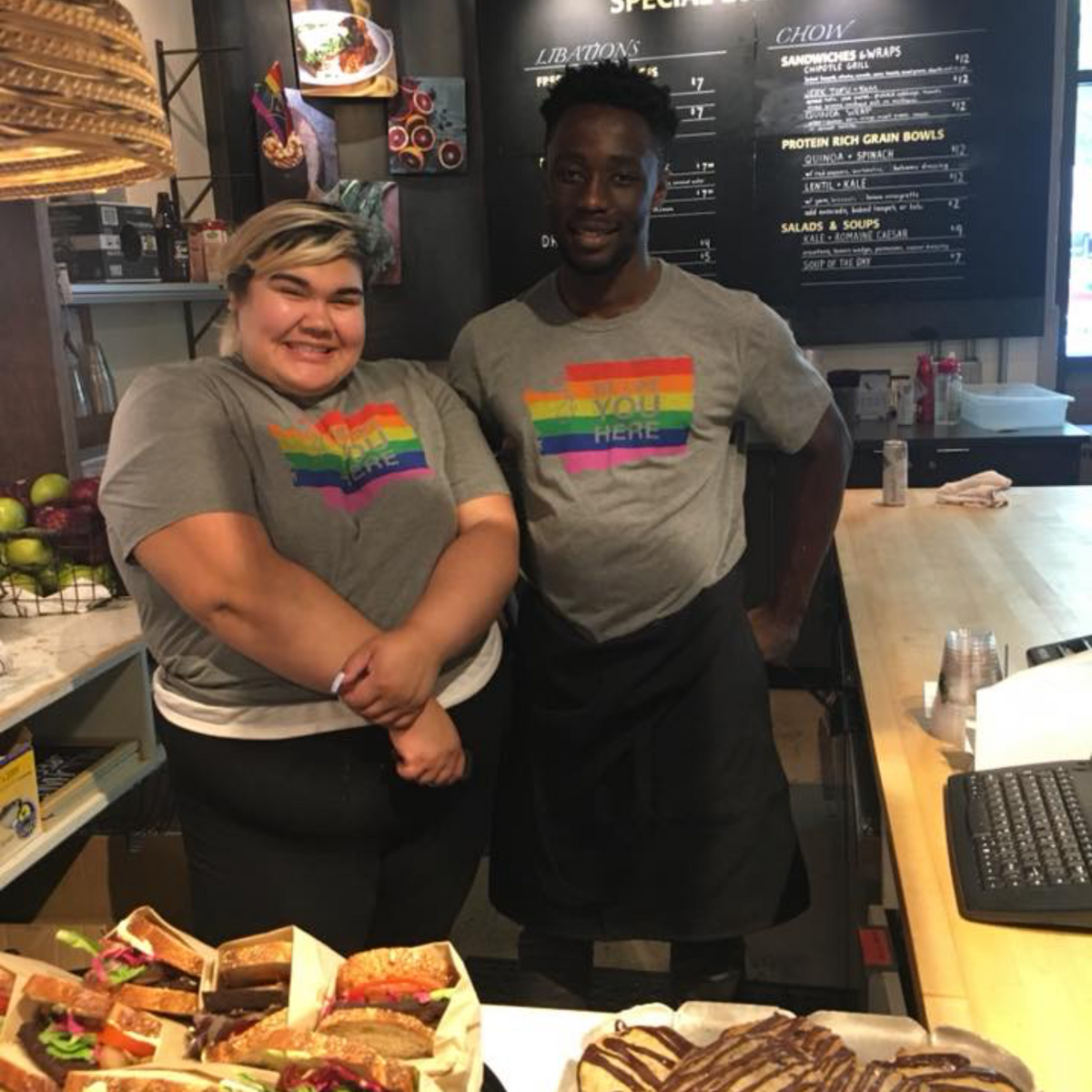 Two people standing behind a counter with sandwiches and pastries, wearing rainbow-themed shirts.