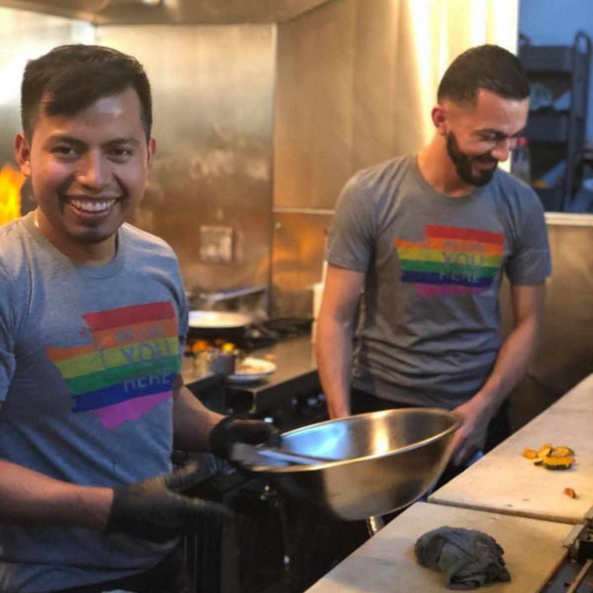 Two men in a kitchen, one wearing a rainbow shirt, preparing food.
