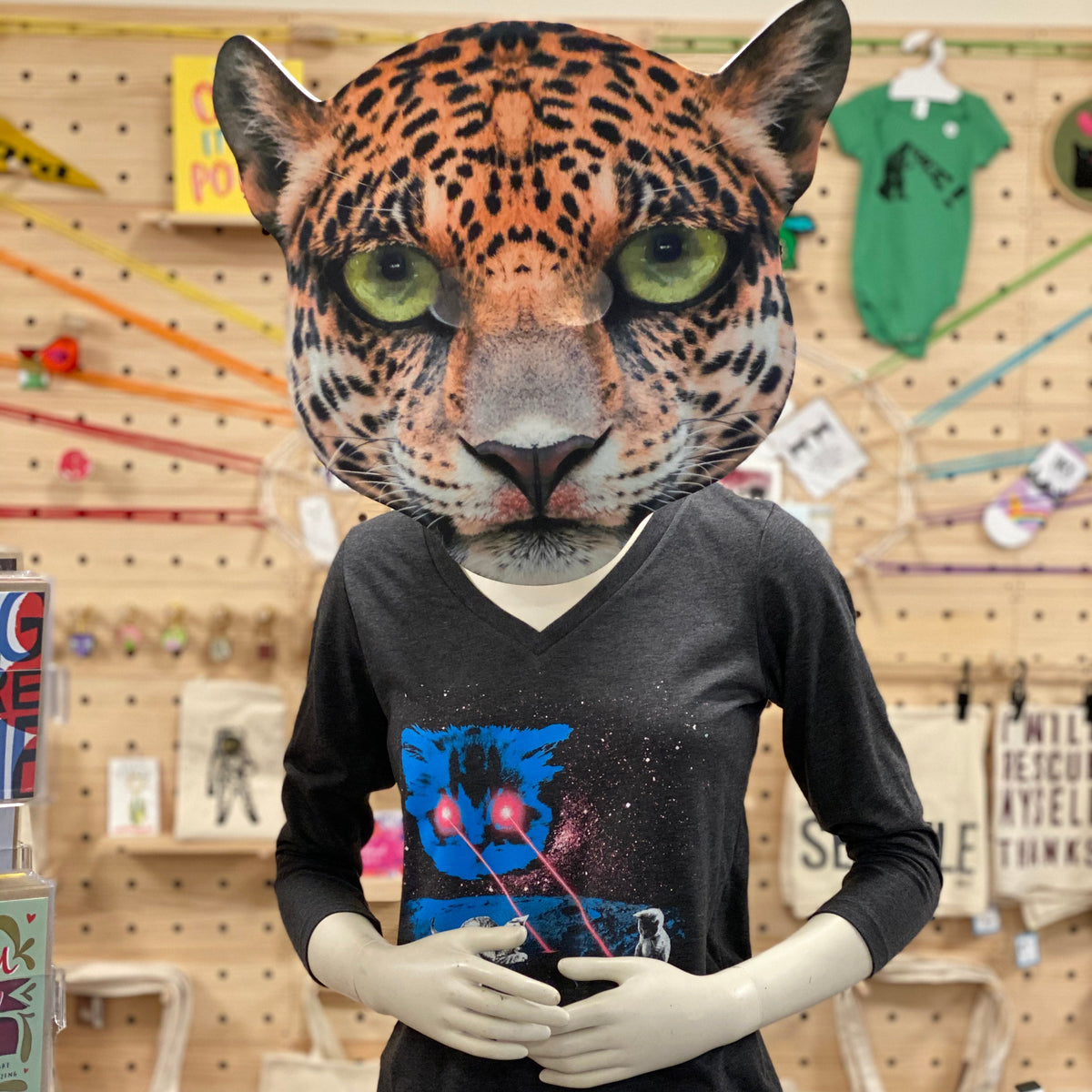Person wearing a leopard mask in a store setting. they are wearing a v-neck long sleeved gray shirt with a space cat on it. The cat is shooting lasers out of its eyes and there are kittens playing with the lasers. 