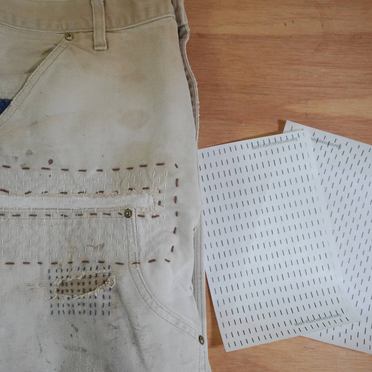 A visibly mended pair of pants next to two stick and stitch patterns against a wooden background. 