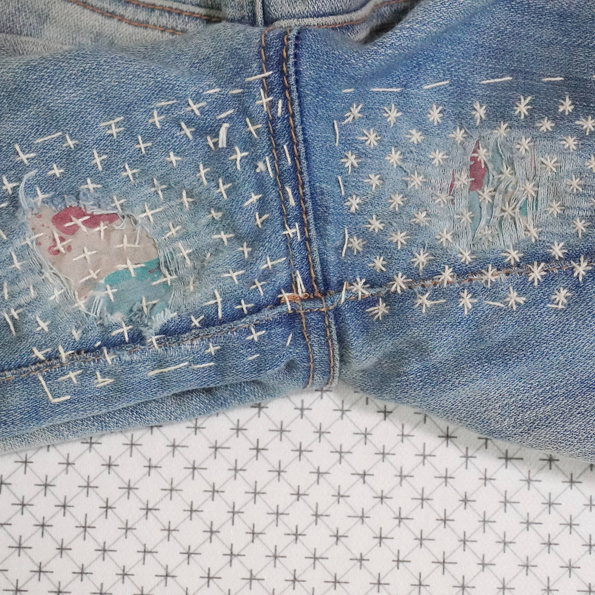 Stitched up example of visible mending on the crotch of a pair of jeans with a stick and stitch pattern in the background. 