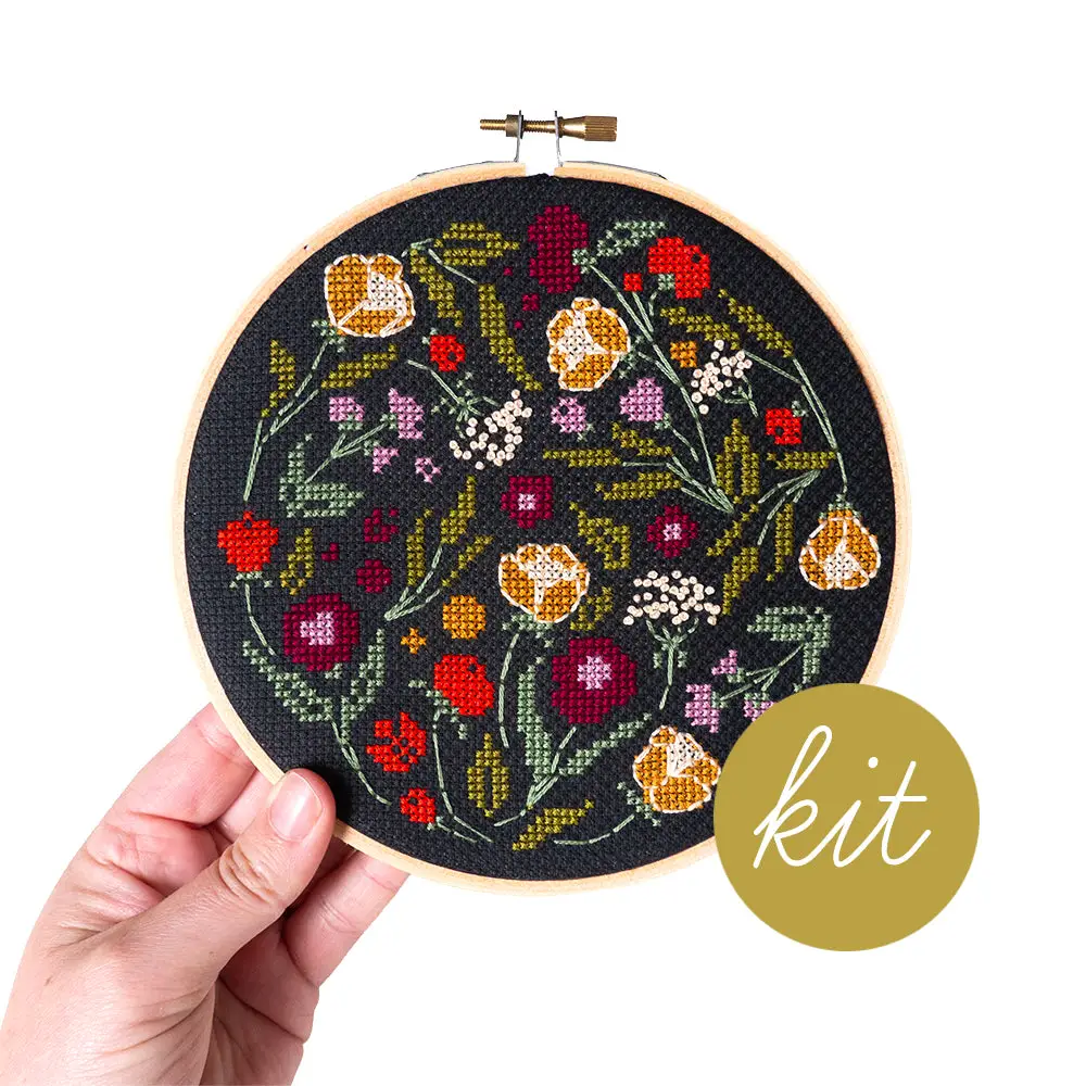 A finished, stitched up sample of the Spring Fling Cross stitch kit by Junebug and Darlin' is being held up by a human hand. The word KIT is added to the front of the photo with an olive green background. 