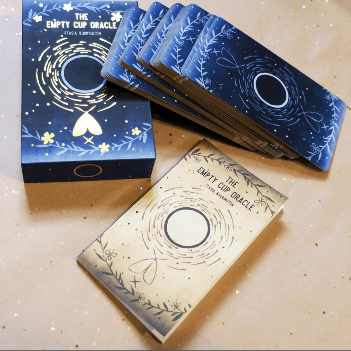 The image shows the 'Empty Cup Oracle Deck' by Stasia Burrington, featuring 40 cardstock cards with minimalist black ink illustrations and gold gilt edging, along with a 60-page guidebook and a sturdy lidded box.