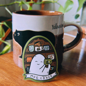 A ceramic mug with a black cat illustration and the word 'Yellowstone' sits on a wooden table, with a die-cut sticker in front of it showing a cute ghost character and the text 'MUG_LOVER'.