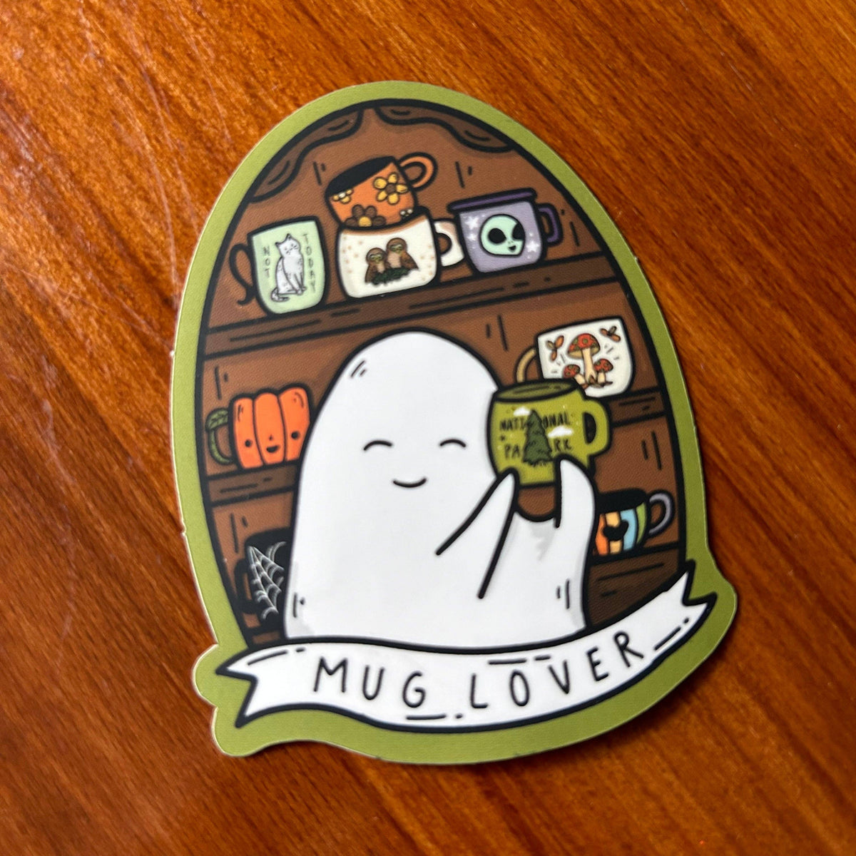 A die-cut sticker featuring a cute white ghost holding a green mug with 'MUG LOVER' written on it, placed on a wooden surface. The sticker has an olive green border and a whimsical coffee shop shelf illustration in the background.