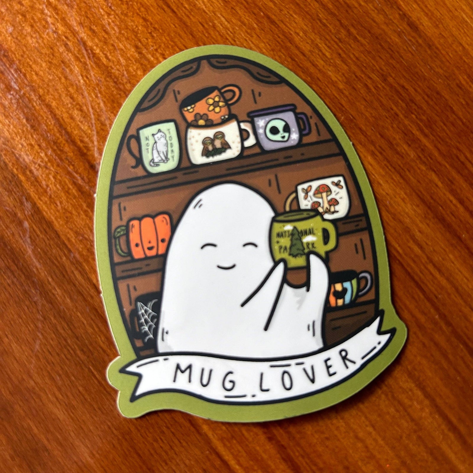 A die-cut sticker featuring a cute white ghost holding a green mug with 'MUG LOVER' written on it, placed on a wooden surface. The sticker has an olive green border and a whimsical coffee shop shelf illustration in the background.