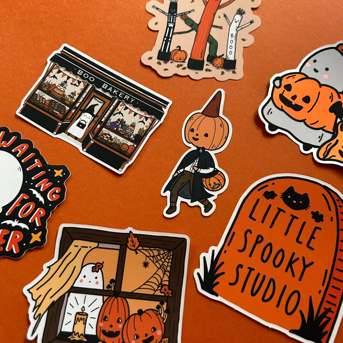 A collection of Halloween-themed die-cut stickers arranged on a solid orange background. The stickers feature cute illustrations including a pumpkin-headed character in a witch outfit, a tombstone reading 'LITTLE SPOOKY STUDIO', a bakery storefront, a window with jack-o'-lanterns and a ghost, and a pumpkin patch scene.