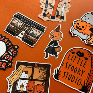 A collection of Halloween-themed die-cut stickers arranged on a solid orange background. The stickers feature cute illustrations including a pumpkin-headed character in a witch outfit, a tombstone reading 'LITTLE SPOOKY STUDIO', a bakery storefront, a window with jack-o'-lanterns and a ghost, and a pumpkin patch scene.