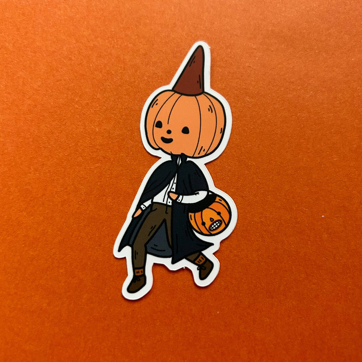 A die-cut sticker featuring a cute cartoon pumpkin character dressed as a vampire, holding a small jack-o'-lantern bucket, shown on a textured orange background.