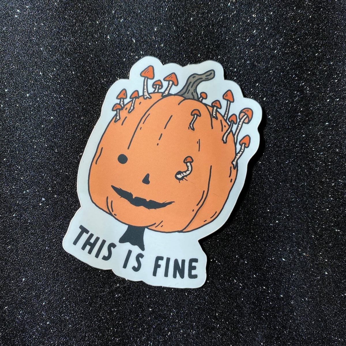 A die-cut sticker featuring a cartoon orange pumpkin with mushrooms growing out of its head and the text 'THIS IS FINE' in black, placed on a dark glittery background.