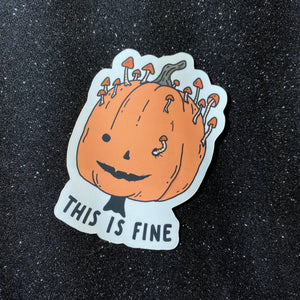 A die-cut sticker featuring a cartoon orange pumpkin with mushrooms growing out of its head and the text 'THIS IS FINE' in black, placed on a dark glittery background.