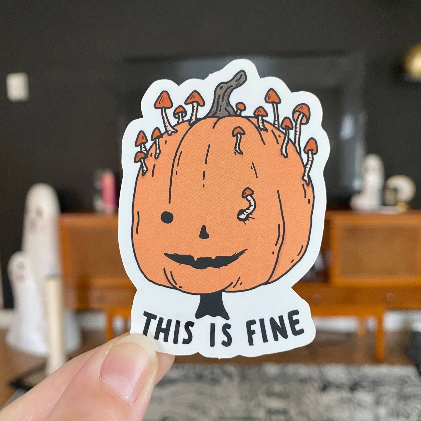 A die-cut sticker held by a hand, featuring a cartoon orange pumpkin with mushrooms growing out of its head and the text 'THIS IS FINE' at the bottom. The background shows a blurred living room interior.