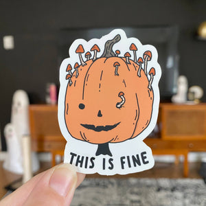 A die-cut sticker held by a hand, featuring a cartoon orange pumpkin with mushrooms growing out of its head and the text 'THIS IS FINE' at the bottom. The background shows a blurred living room interior.