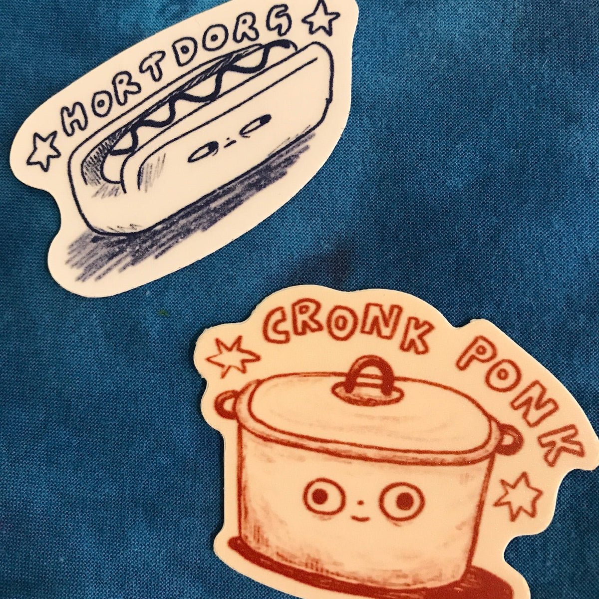 Two die-cut stickers displayed on a textured blue fabric background. The top sticker features a cute hand-drawn hot dog with the text 'HORTDORG' in black ink. The bottom sticker is a crock pot with a face and it says 'CRONK PONK" 