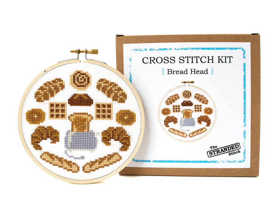 A cross stitch kit with a design that resembles a loaf of bread and various baked goods, displayed in an embroidery hoop. It is standing next to the brown kraft box that the kit comes in.