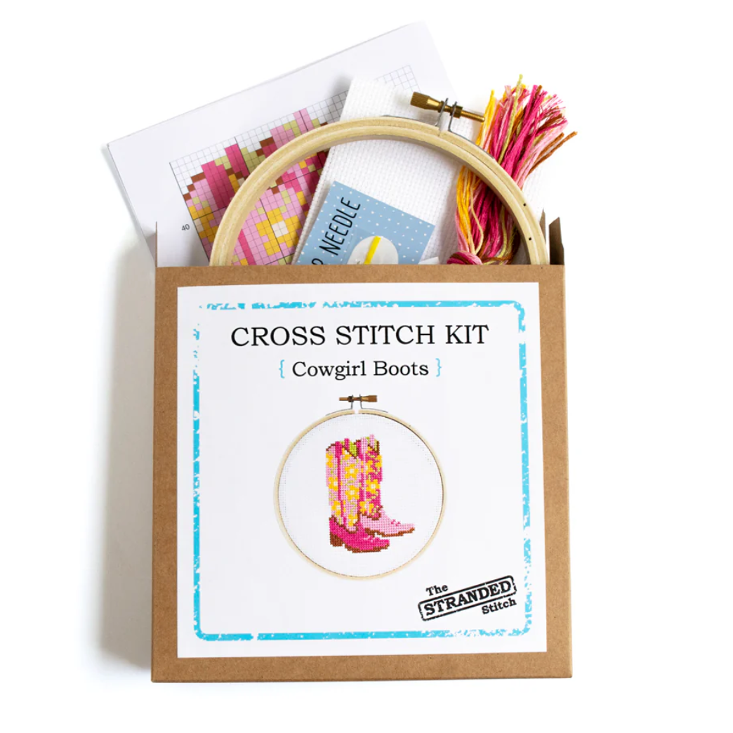 Cross stitch kit with pink cowgirl boots design on a white background. This photo is of the brown kraft box packaging with all the supplies coming out of the top. 