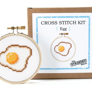 A completed cross stitch depicting a fried egg, mounted in a 3 inch embroidery hoop. It is standing in front of the brown kraft box that the kit comes in.