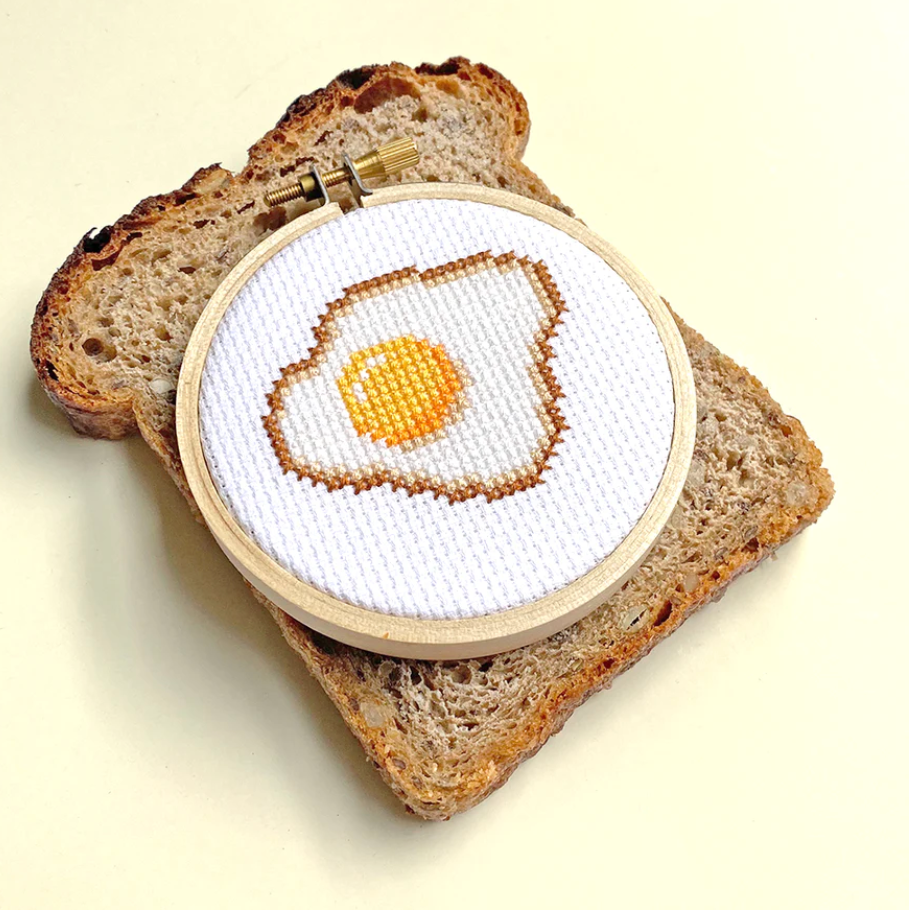 A completed cross stitch depicting a fried egg, mounted in a 3 inch embroidery hoop. It is set atop a piece of wheat toast.