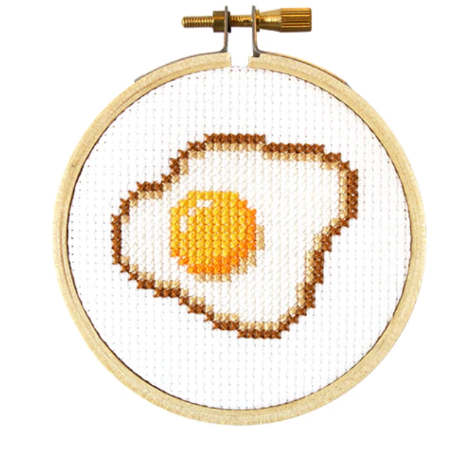 A completed cross stitch depicting a fried egg, mounted in a 3 inch embroidery hoop.