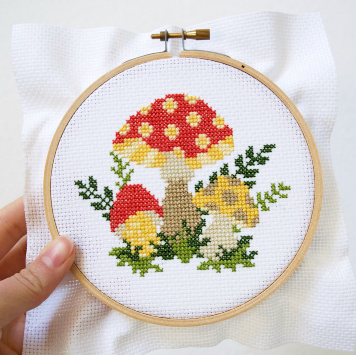 A hand holding a cross stitch kit with a mushroom design, featuring a white aida cloth, embroidery hoop, and colored threads.