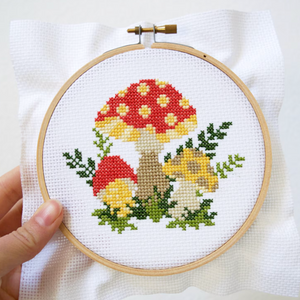 A hand holding a cross stitch kit with a mushroom design, featuring a white aida cloth, embroidery hoop, and colored threads.