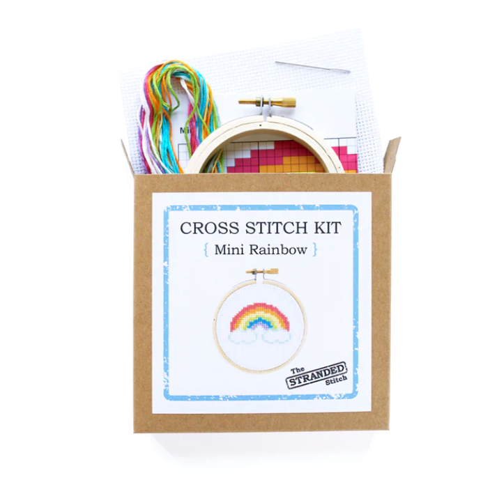 A brown kraft box with a sticker showing a completed mini rainbow cross stitch kit. The supplies included in the kit are coming out the top. 