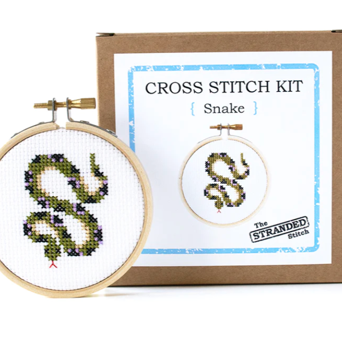 A completed snake cross stitch kit in a three inch hoop. It stands in front of the kraft brown package that the kit comes in. 