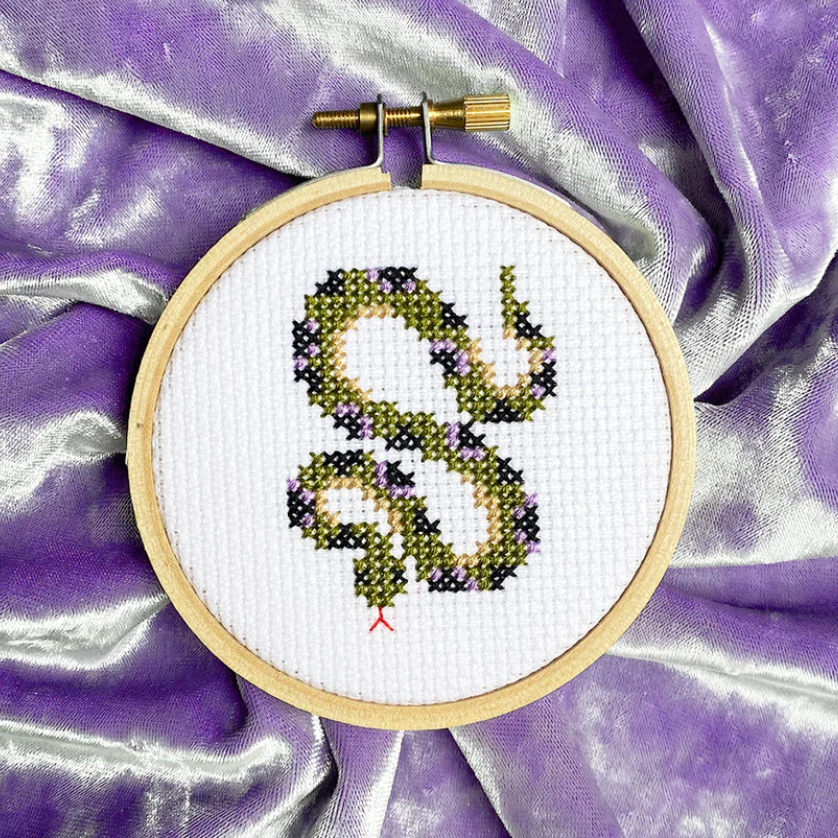 A completed mini snake cross stitch in a three inch hoop. It is laying on a piece of lavender velvet. 