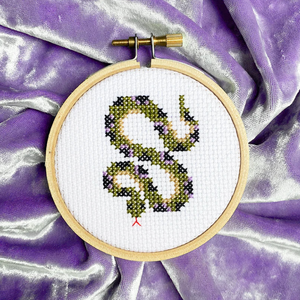A completed mini snake cross stitch in a three inch hoop. It is laying on a piece of lavender velvet. 