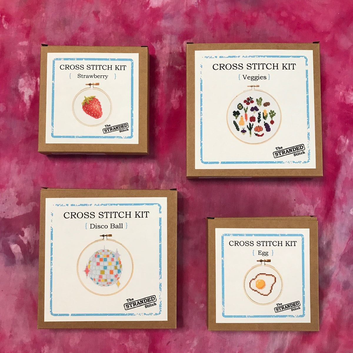 A set of four cross stitch kits laid out on a pink ice dyed tablecloth: Strawberry, Veggies, Disco Ball & Egg.