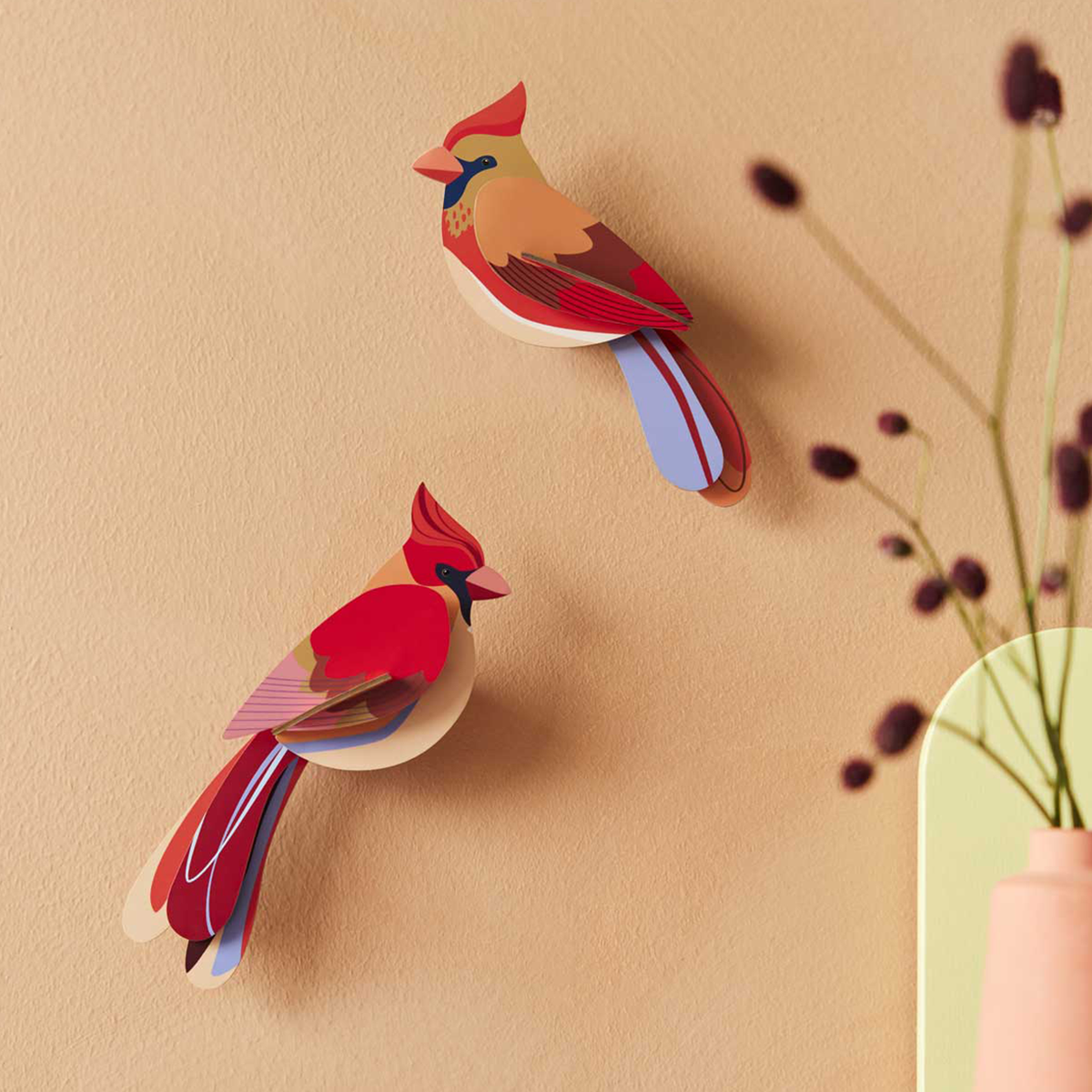 Two paper cardinal sculptures hanging on a peach colored wall with dried flowers in the foreground. 