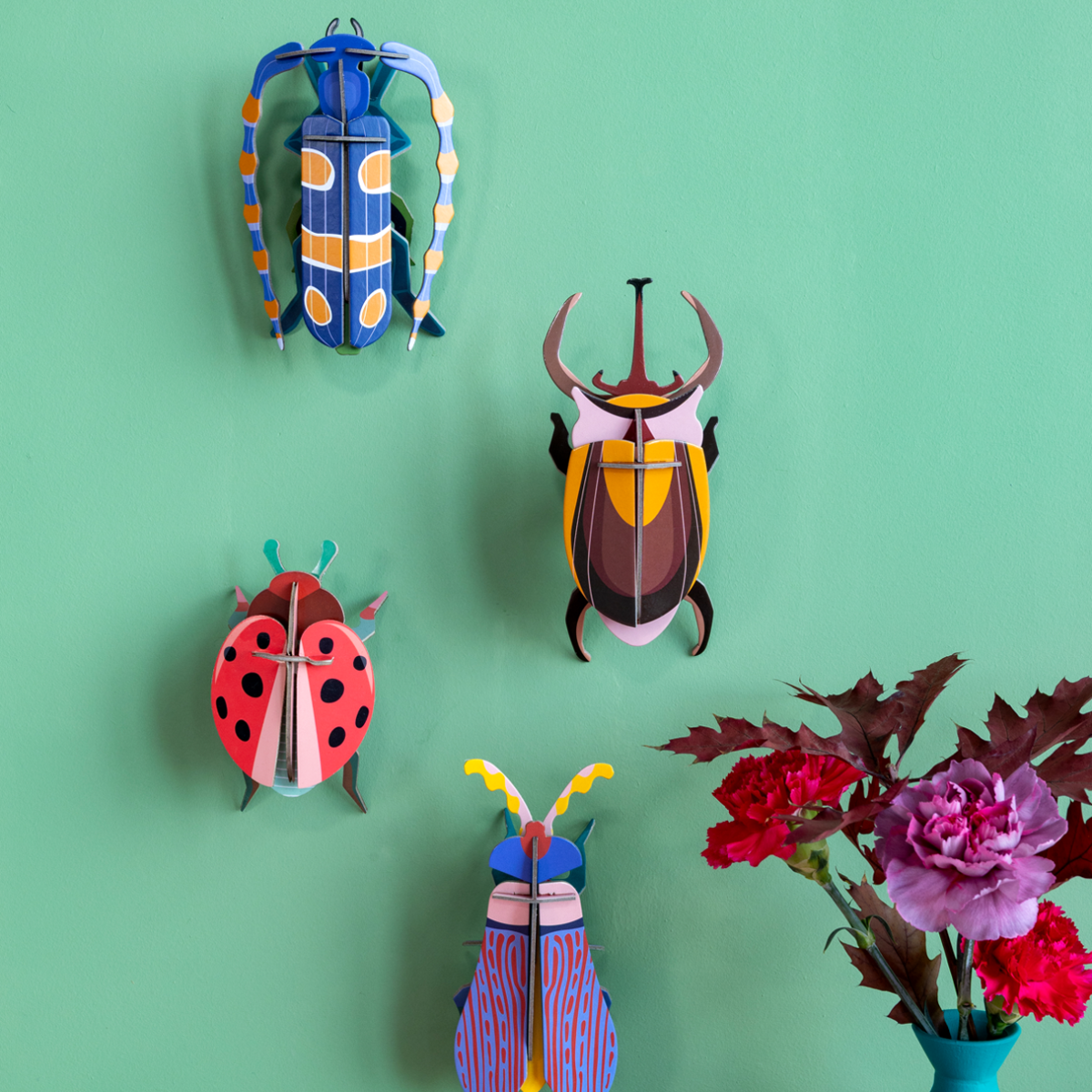 Grouping of bug-themed cardboard wall art sculptures hanging on a bright teal wall with flowers in the foreground. 