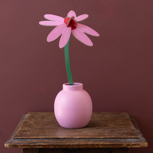 A pink and red flower made out of cardboard in a pink vase on a wooden table against a dark brown background. 