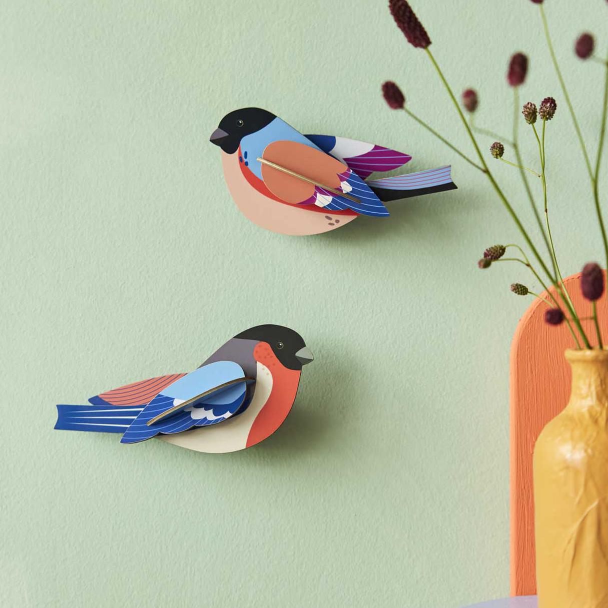 Two cardboard finch wall art pieces on a light green wall with dried flowers in a yellow vase in the foreground. 