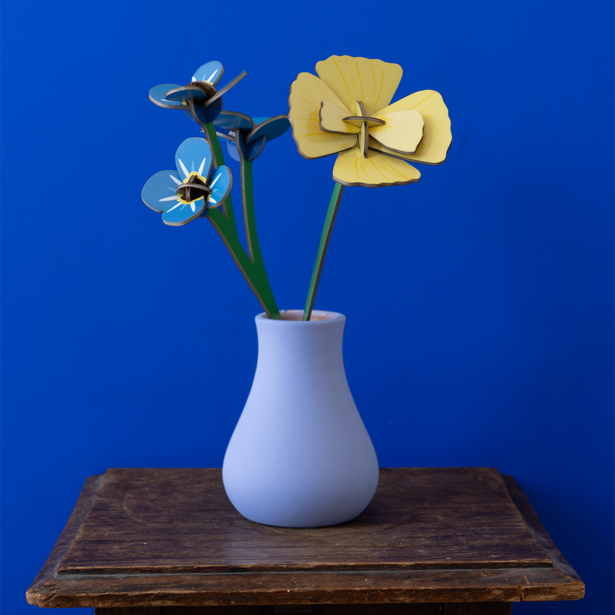 Cardboard flowers in a light blue vase on a wooden table against a bright blue wall. 