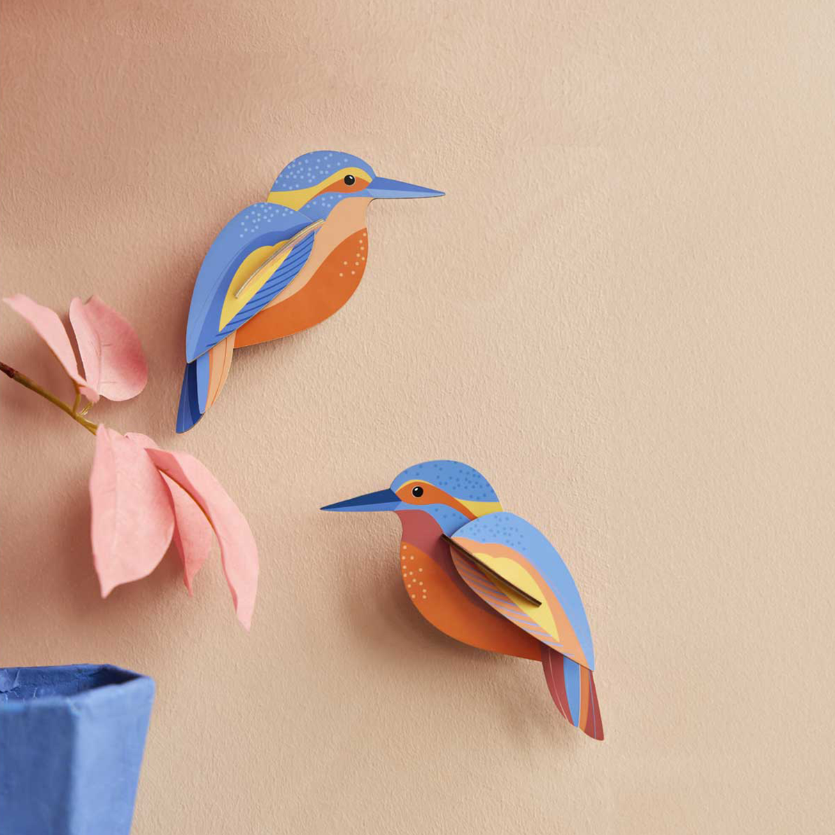 Two colorful cardboard sculptures of kingfishers mounted on a peach colored wall. 