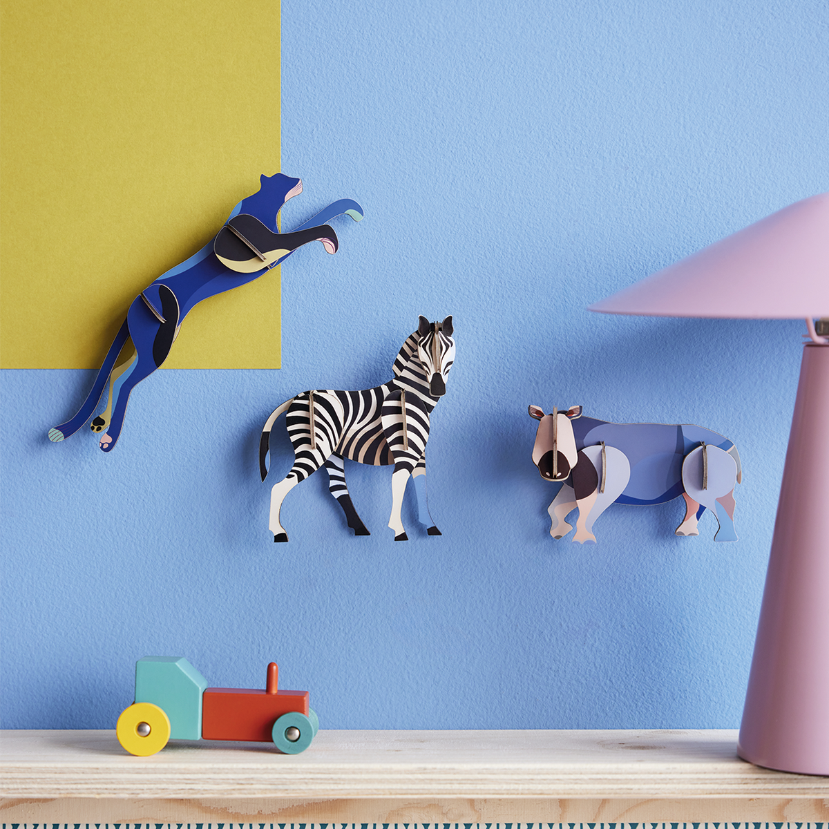 Paper sculptures of a panther, zebra, and rhino mounted on a periwinkle wall with a wooden toy train in the foreground. 
